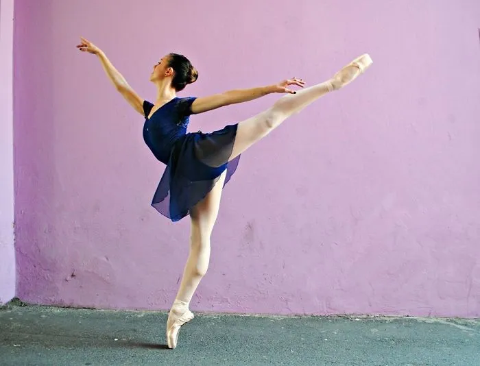 Ballerina in a blue dress performing an arabesque against a purple wall for Ballet Day.