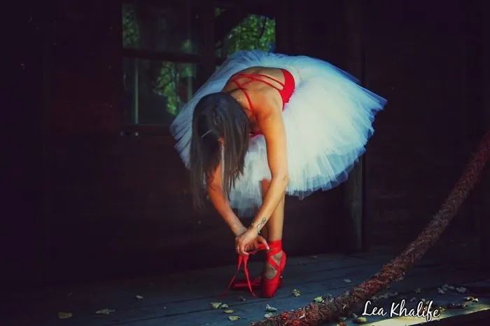 Ballet dancer in a white tutu adjusting red pointe shoes in a dimly lit studio.