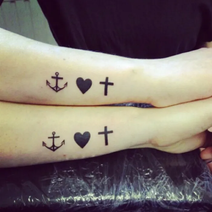 Two arms with matching sister tattoos featuring an anchor, heart, and cross design, symbolizing a strong bond.