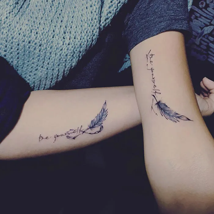 Matching sister tattoos on arms with feathers and "be yourself" text.