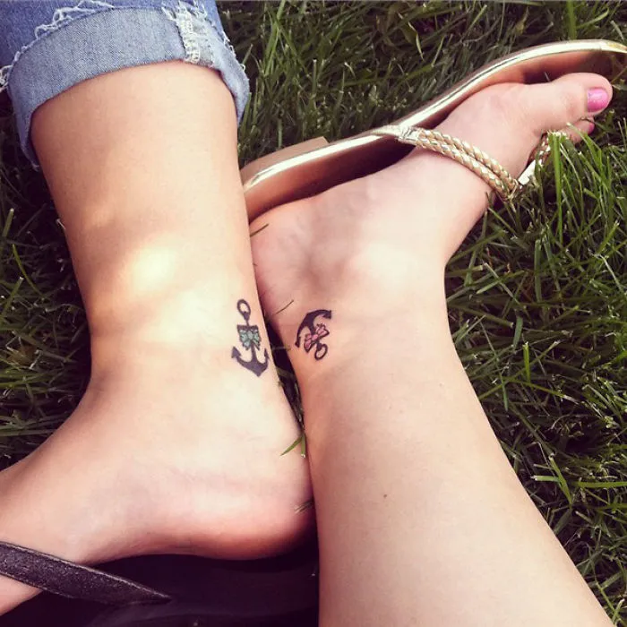 Feet with matching sister tattoos featuring an anchor and a rose on grass.