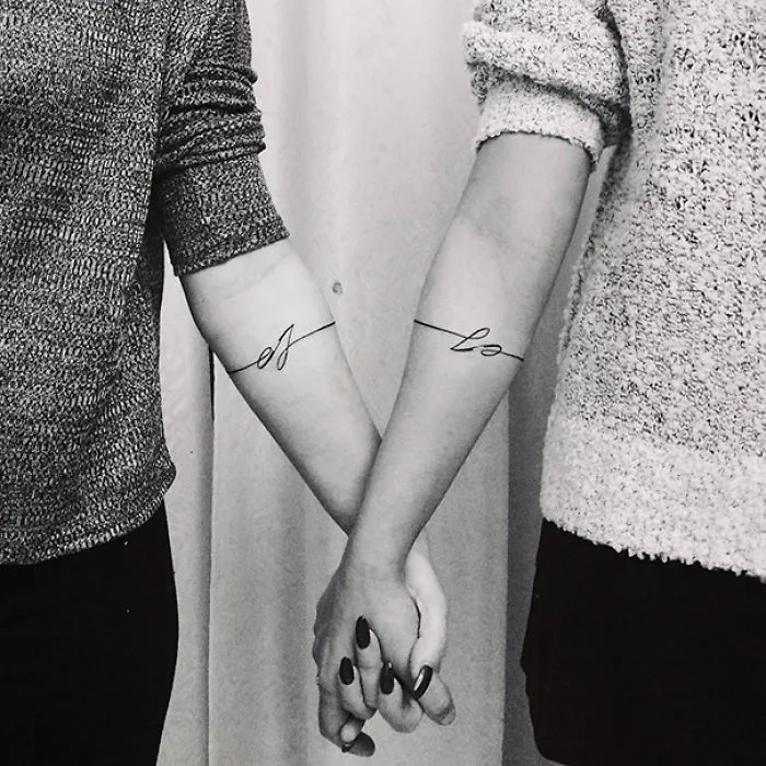 Sisters holding hands, showcasing minimalistic tattoos on forearms symbolizing their bond.