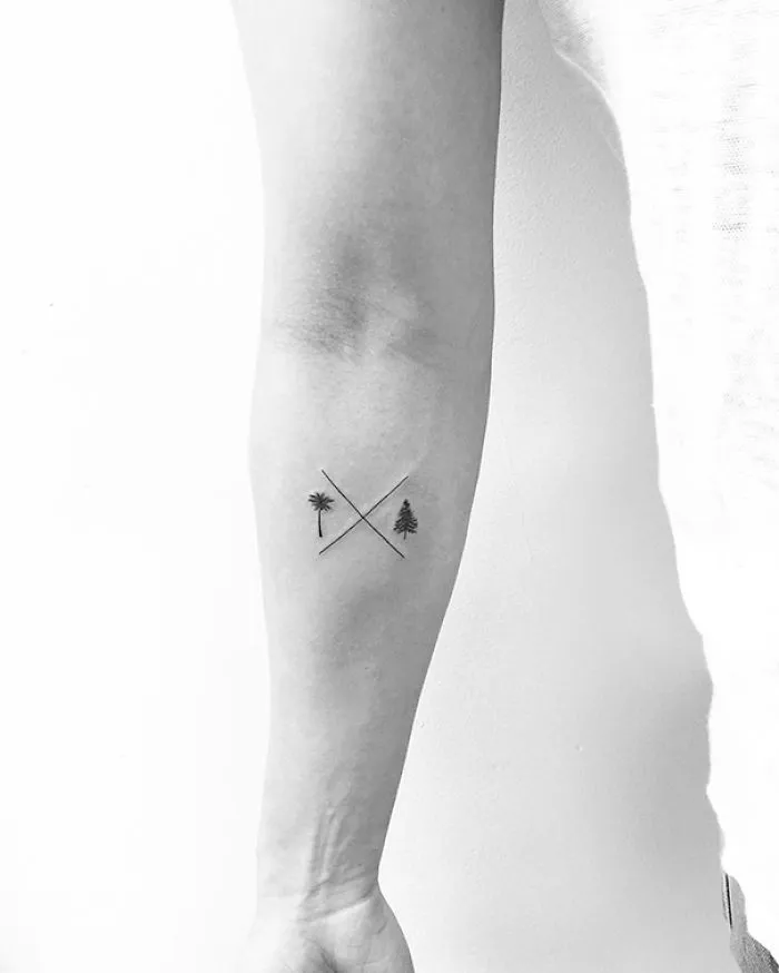 Minimalist tattoo of crossed lines with palm and pine trees on forearm.