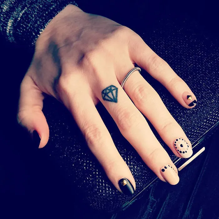 Minimalist tattoo of a diamond on a finger, hand resting on a textured surface with black and white nail art.