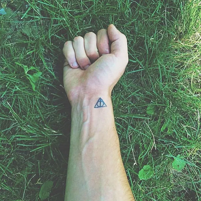 Minimalist tattoo on wrist with a geometric symbol, displayed on a grass background.