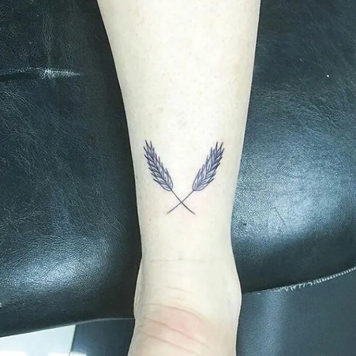Minimalist tattoo of two crossed wheat stalks on an arm, showcasing simple inked designs.