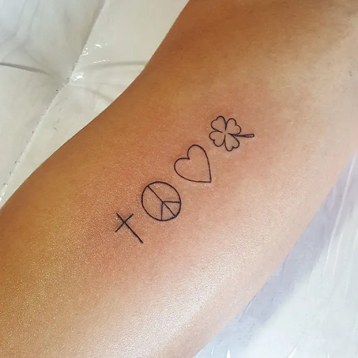 Minimalist tattoo with symbols: cross, peace sign, heart, and clover on forearm.