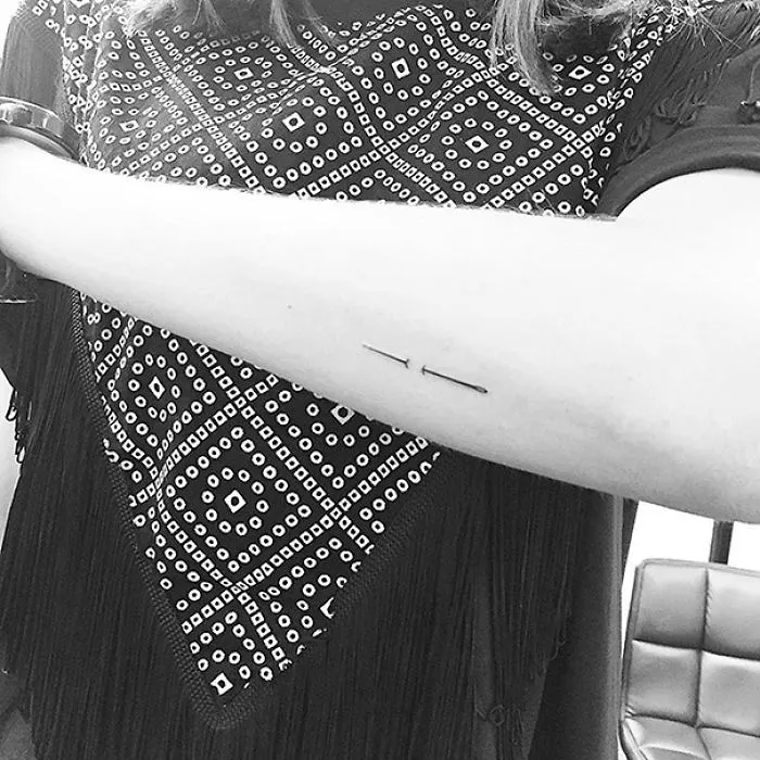 Minimalist tattoo of two thin lines on forearm against patterned shirt background.