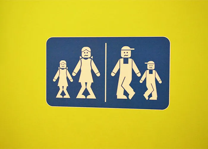 Funny bathroom sign with stick figures, woman and daughter on left, man and son on right, on a yellow background.