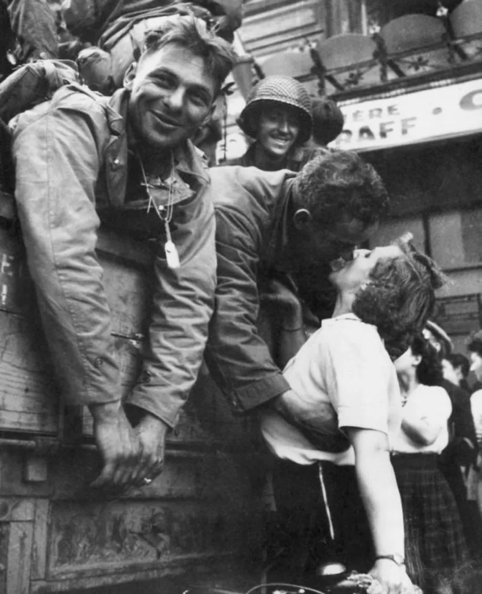 Soldier kissing a woman goodbye during wartime, with another soldier smiling by their side, capturing a moment of love.