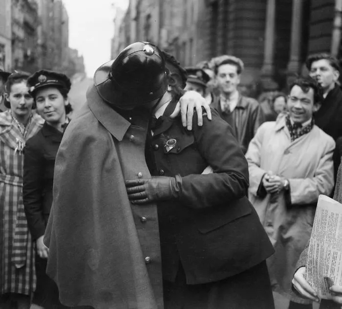 Soldier and woman embrace passionately in a historic wartime street scene, surrounded by onlookers.