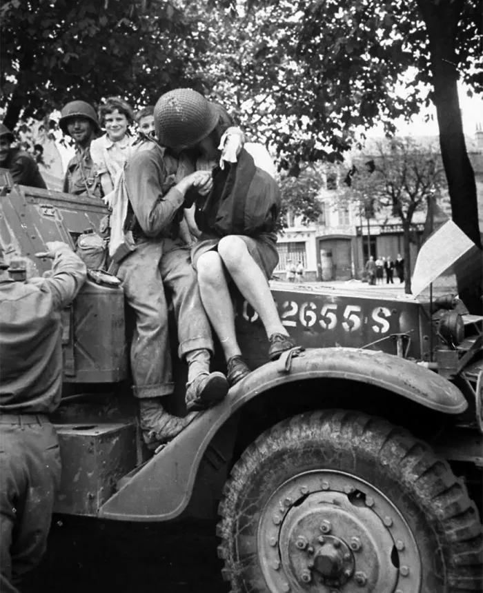 Soldier and woman kissing on a wartime vehicle, surrounded by smiling troops under a tree.