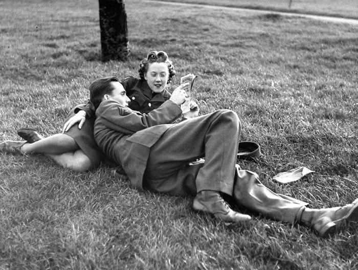 Couple in military attire lying on grass, reading together, depicting love during wartime.
