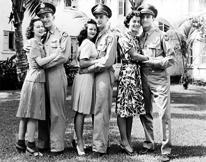 Couples embracing, showcasing love during wartime, with soldiers in uniform and women in dresses, outside a building.