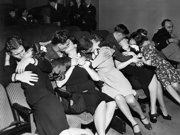 Couples embracing during wartime, seated in a row at a theater, showcasing love amid conflict.