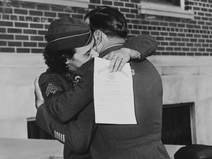 Historic photo of a soldier embracing a woman during wartime, holding a document, against a brick building backdrop.