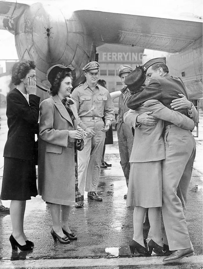 Couple embraces during wartime near an aircraft, surrounded by smiling onlookers, showcasing historic love moments.