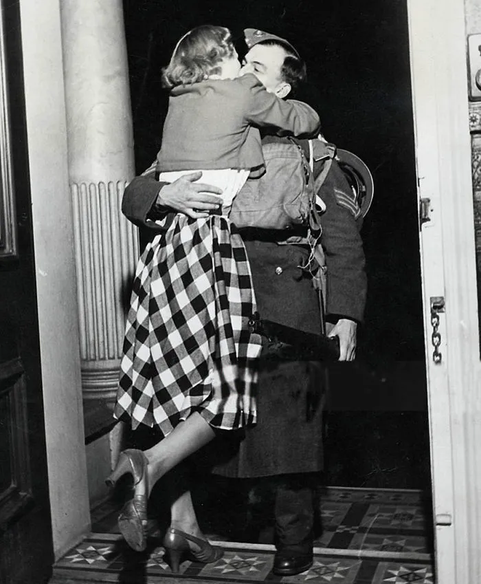Wartime love captured as a soldier embraces a woman in checkered dress, sharing a kiss at the doorstep.