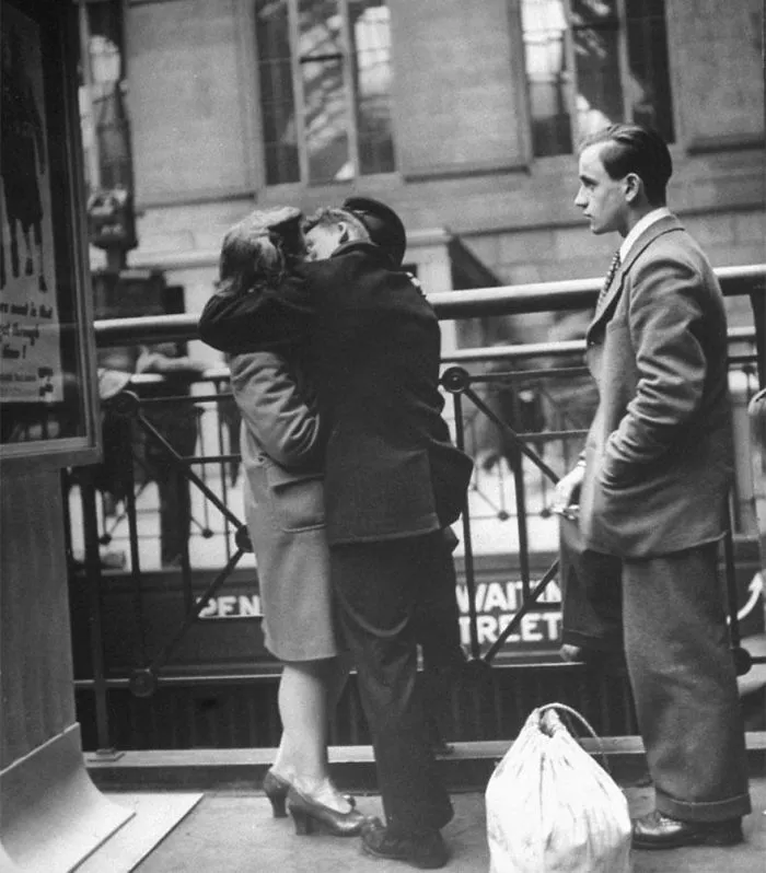 Couple embracing at a train station during wartime, with another man waiting nearby, capturing historic love moments.