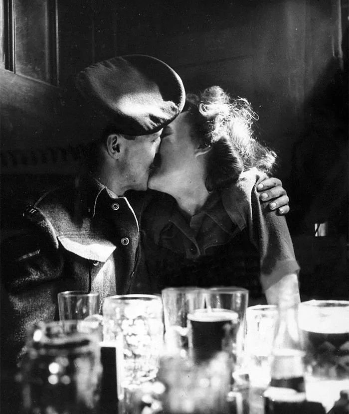 Vintage wartime photo of a soldier and woman sharing a kiss, surrounded by glasses.
