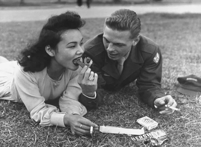 Couple enjoying chocolate on grass during wartime, sharing a tender moment.