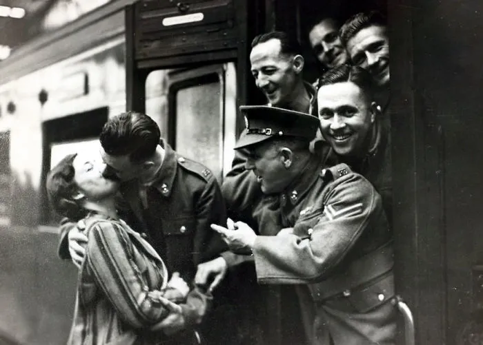Soldier kissing a woman goodbye at a train during wartime, surrounded by smiling comrades leaning out of the train window.