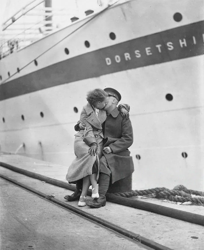 Couple sharing a kiss on a dock during wartime, with a large ship in the background.