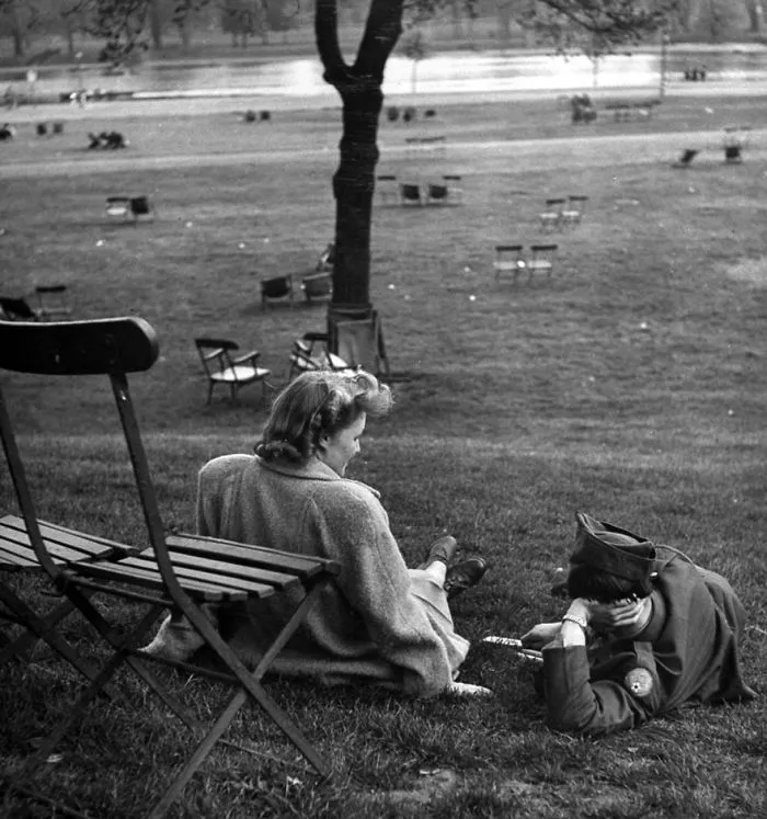 Couple sitting on grass shows love during wartime, with scattered chairs and a tree in the background.