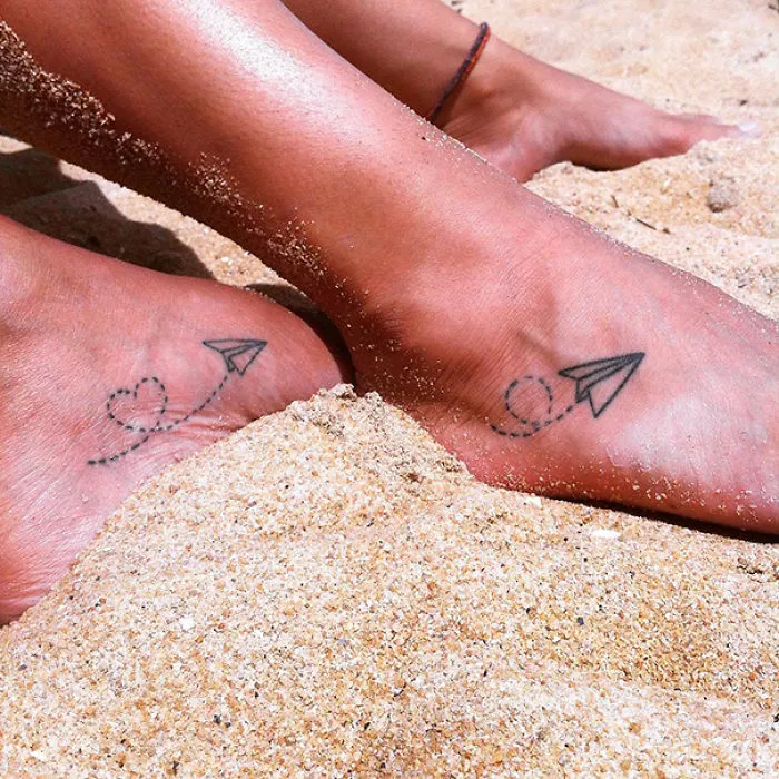 Simple small foot tattoo with a paper plane design on sandy feet.