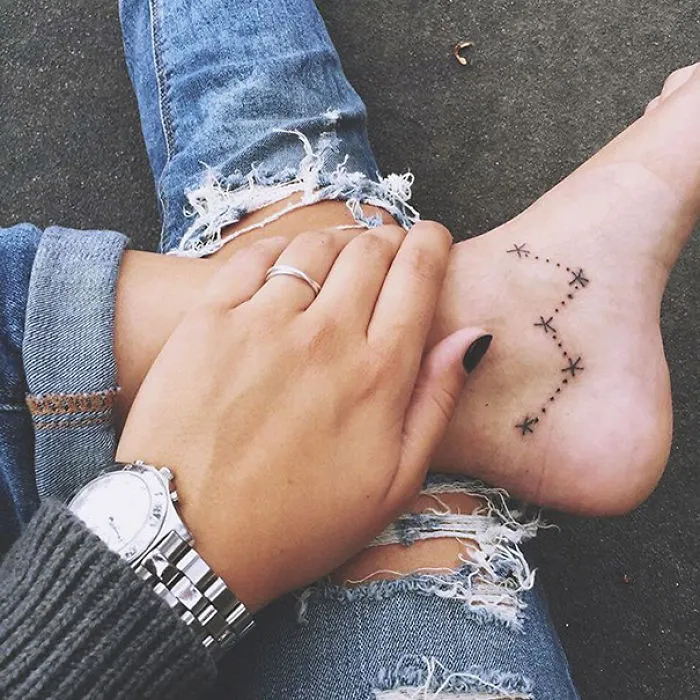 Small foot tattoo of a constellation symbol, worn with ripped jeans and a silver watch.