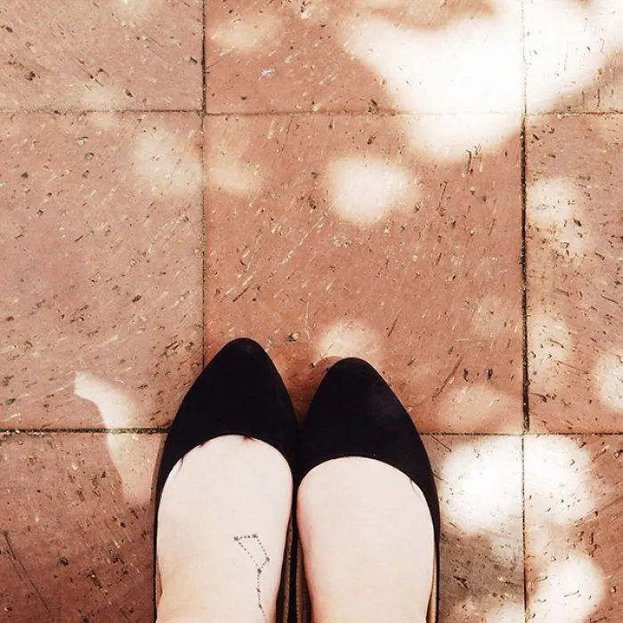 Minimalist small foot tattoo with black shoes on a sunlit brick floor.