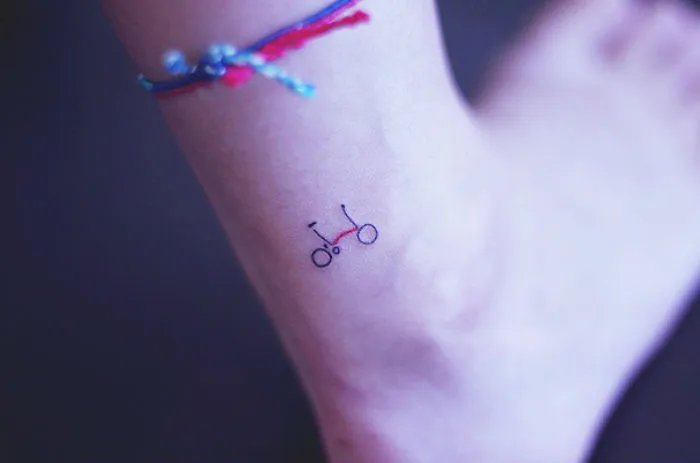 Small foot tattoo of a minimalist bicycle with a colorful ankle bracelet.