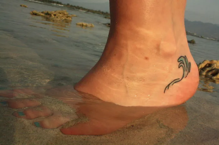 Small foot tattoo of a wave, partially submerged in water, showcasing minimalist design.