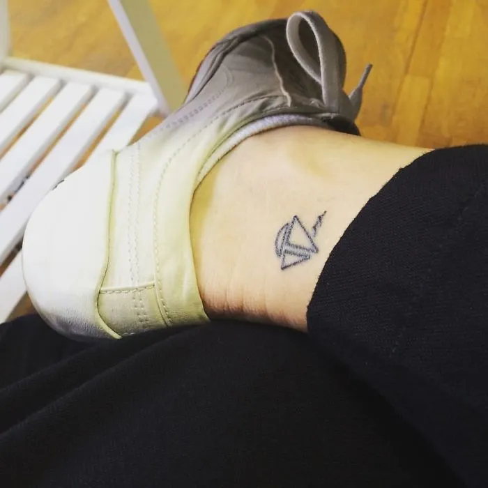 Minimalist small foot tattoo of a paper airplane on the ankle, emphasizing that less is more in tattoo design.