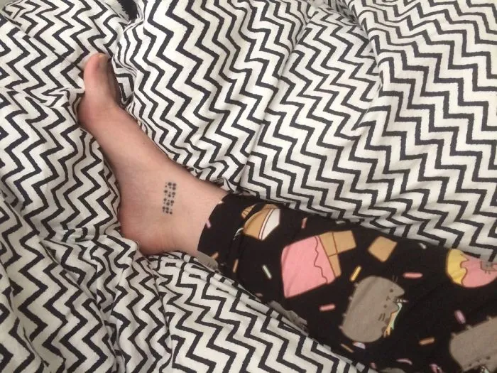 Small foot tattoo on ankle surrounded by patterned sheets, showcasing minimalist ink design.