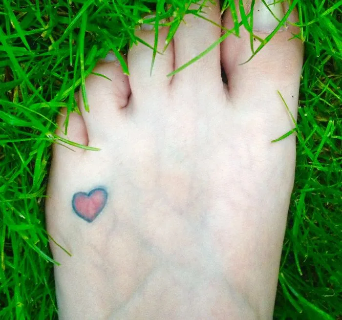 Small foot tattoo of a heart design surrounded by green grass, showcasing minimalist style.