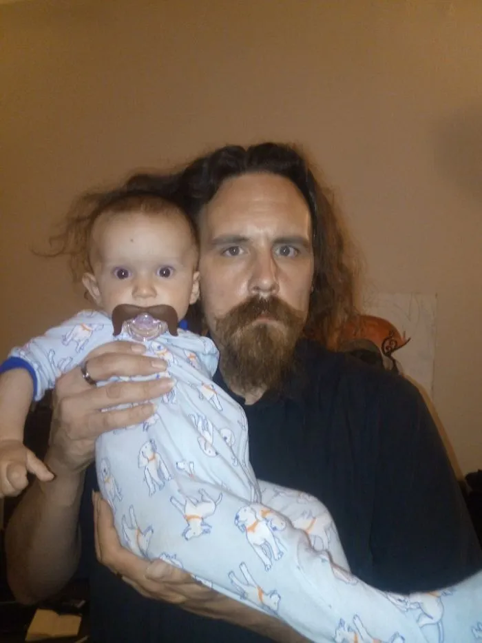 Dad holding a baby with a pacifier shaped like a mustache, funny kids can't be left alone with their dads