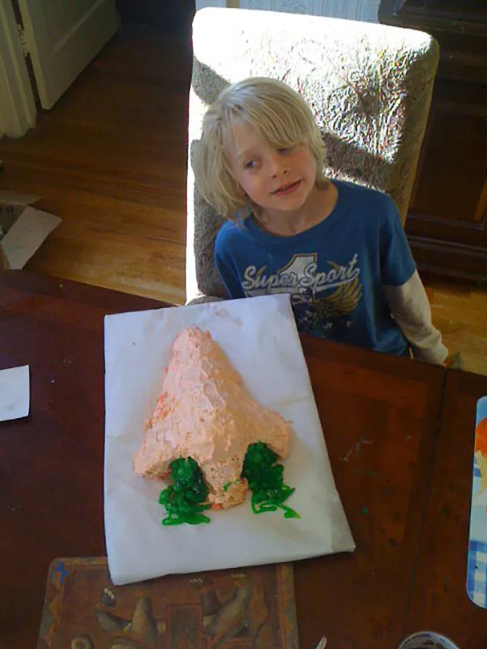 Blond child at table with a cake shaped like a nose and green icing kids alone with dads