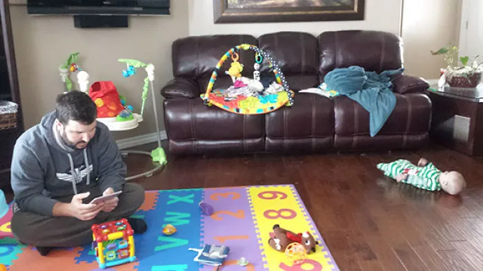 Dad distracted on phone while baby lies on floor play mat