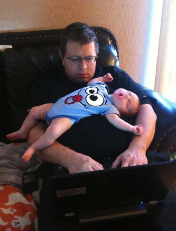 Dad holding sleeping baby while using laptop showing kids left alone with dads