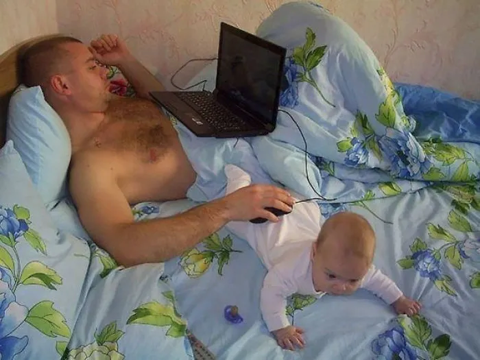 Dad watching laptop in bed with baby crawling next to him showing kids left alone with dads