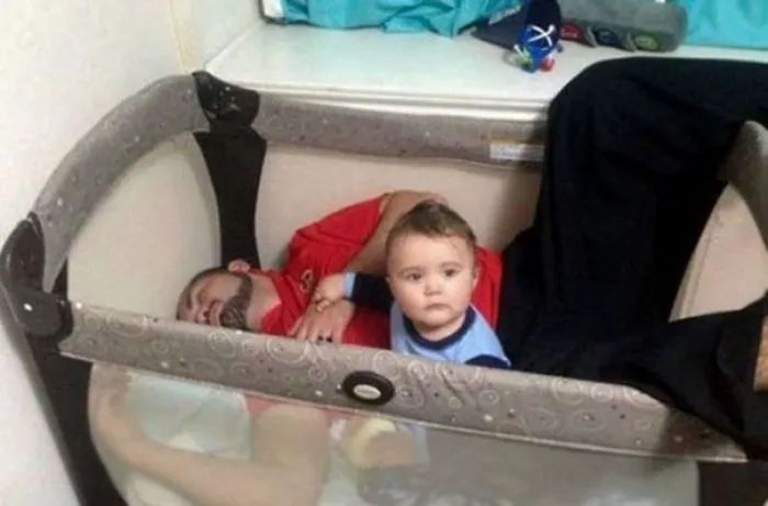 Dad sleeping inside a baby playpen with baby sitting