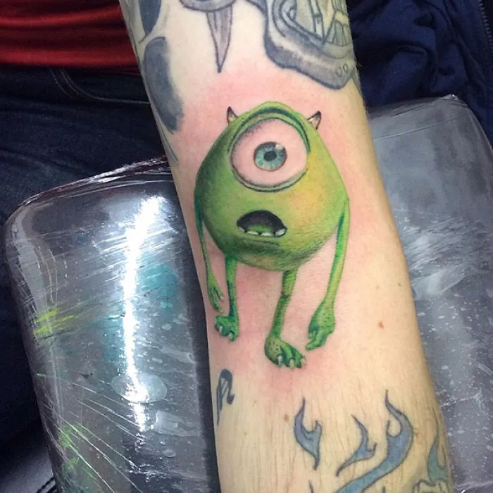 Pixar-inspired tattoo of a green monster with one eye on a forearm.