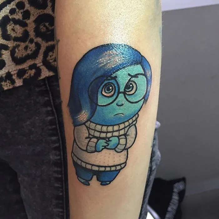 Pixar-inspired tattoo of Sadness from "Inside Out" on an arm, featuring blue hair and glasses.