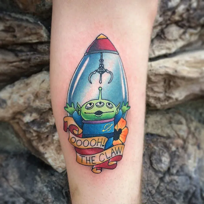 Pixar-inspired tattoo of a green alien under a claw machine, with a banner reading "OOooh! The Claw."