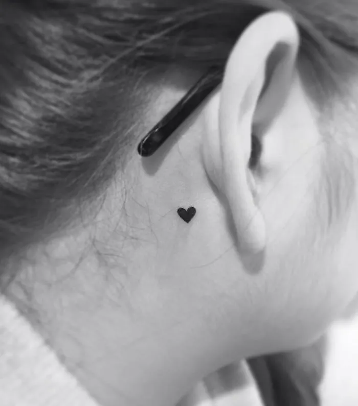Minimalist heart tattoo behind ear by a Korean artist, showcasing simple and elegant design.