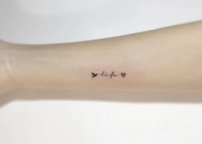 Minimalist tattoo on arm featuring the word "life" with a small bird and heart design.