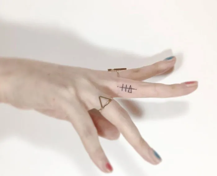 Minimalist tattoo of geometric symbols on a finger by a Korean artist, hand adorned with rings, against a light background.