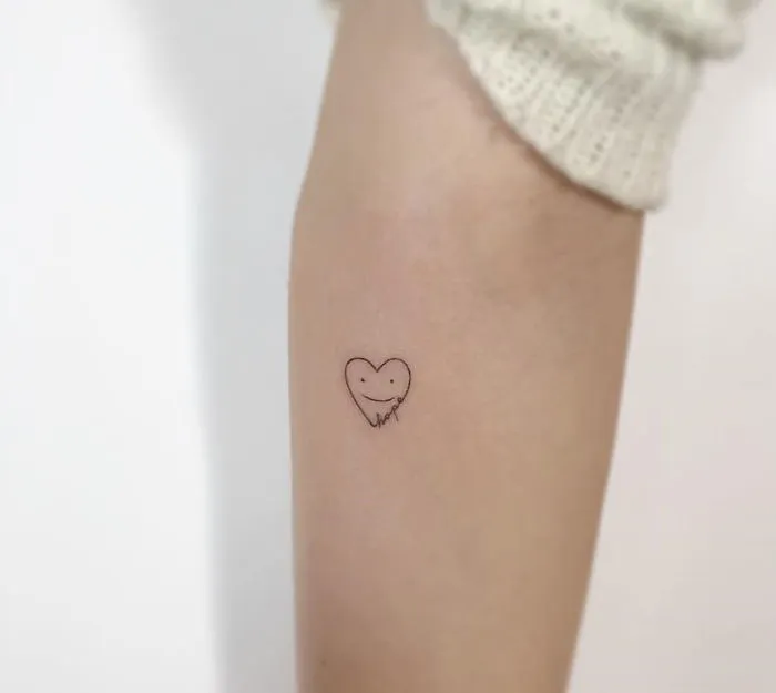 Minimalist tattoo of a heart with a smiley face on an arm, designed by a Korean artist.