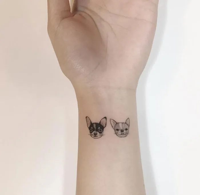 Minimalist tattoos featuring two small dog faces on a person's wrist by a Korean artist.